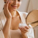 Mulher aplicando creme facial em um ambiente aconchegante com luz suave, rodeada por produtos de skincare organizados em uma bancada de madeira clara.