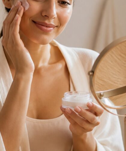 Mulher aplicando creme facial em um ambiente aconchegante com luz suave, rodeada por produtos de skincare organizados em uma bancada de madeira clara.